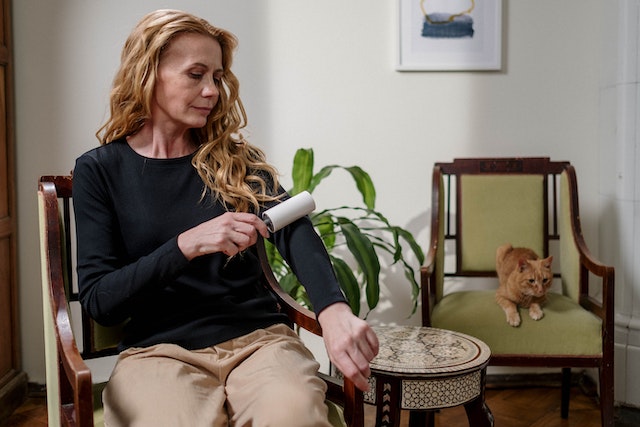 Photo of a woman sitting in a chair and removing pet hair from her clothes using a roller and her orange cat sitting on a chair next to her.