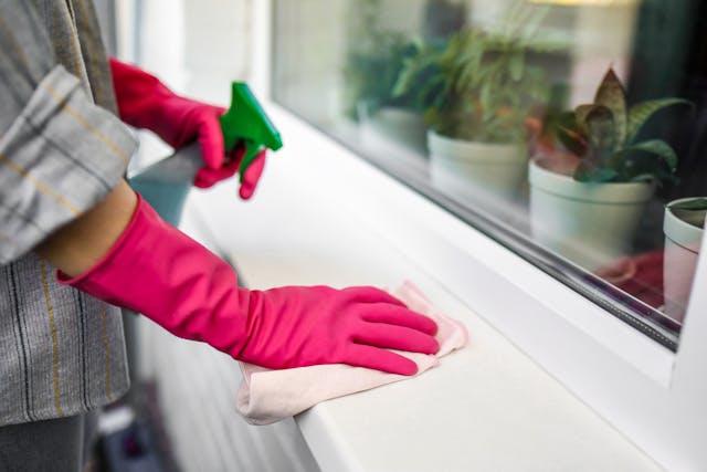 A woman in pink gloves holding a green cleaning product bottle cleaning a white surface.